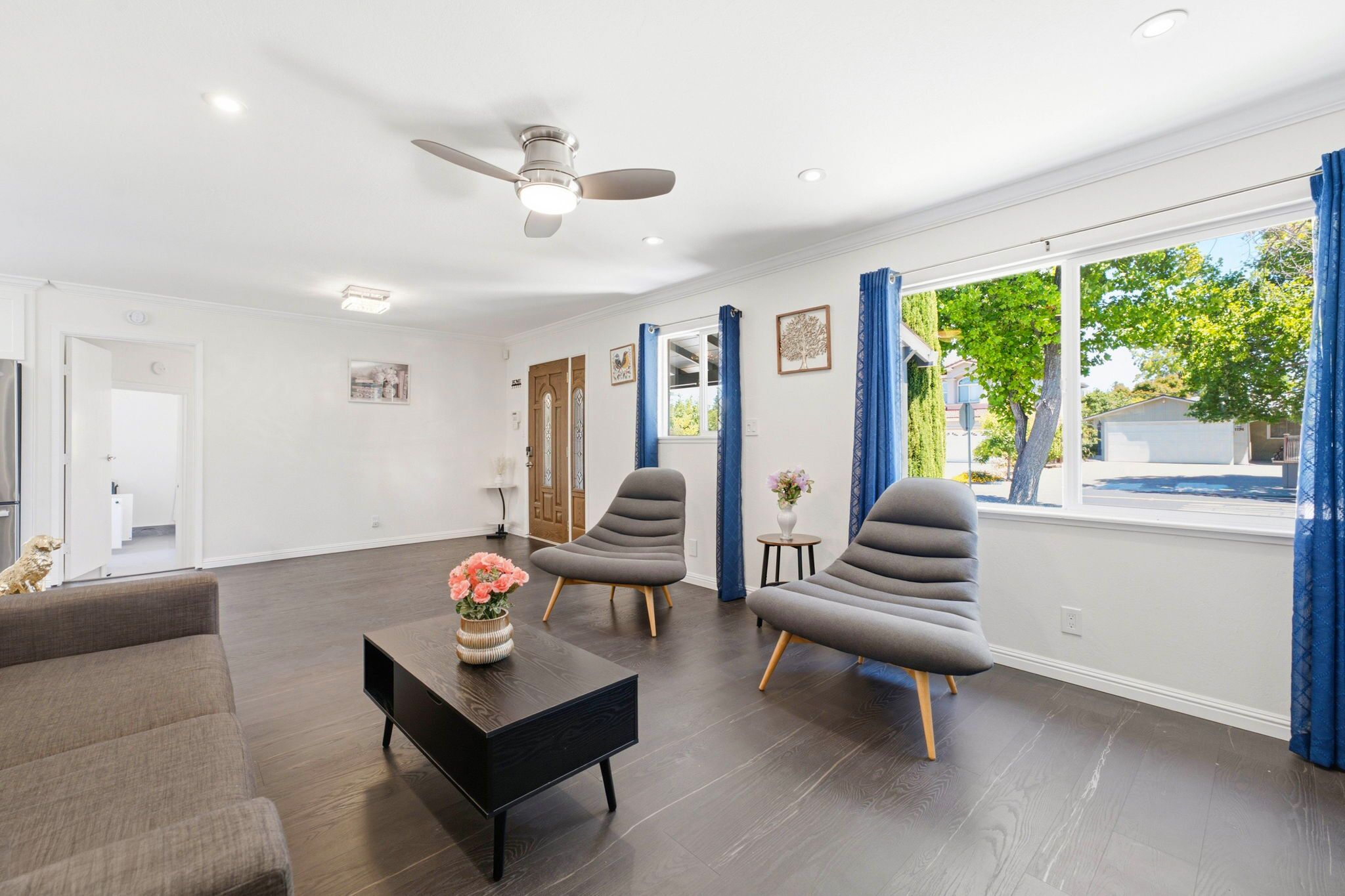 Un salon moderne avec un parquet en bois foncé, un ventilateur de plafond et une grande fenêtre ornée de rideaux bleus.