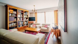 Ein Wohnzimmer mit einer Couch, Sesseln, einem Couchtisch, einem Bücherregal, einem Fernseher und einem Bild an der Wand.