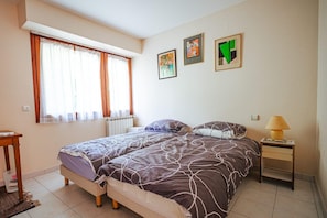 A bedroom with a bed, two pillows, a bedside table with a lamp, a radiator, and a window with curtains. - L'Orée du Bois - SAUGNAC ET CAMBRAN (SAUGNAC ET CAMBRAN)