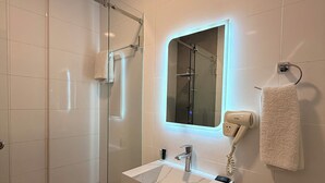 Un baño moderno con ducha envuelta en vidrio, un lavabo blanco con encimera de mármol y un secador de pelo fijado a la pared.