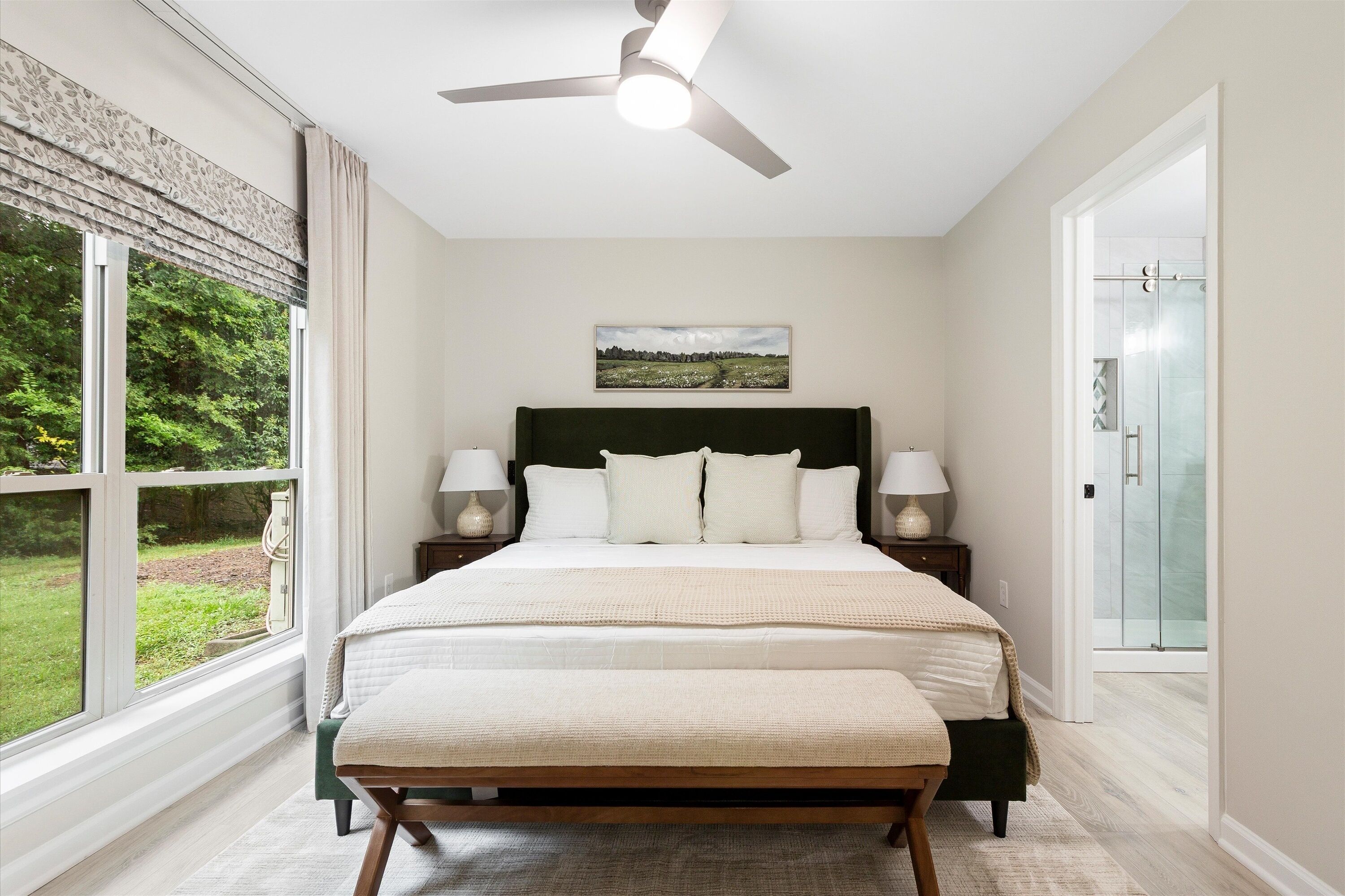 Une chambre à coucher comprenant un lit, des lampes de chevet, un ventilateur de plafond, une fenêtre avec vue et une porte donnant sur une salle de bain.