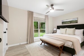 A bedroom with a bed, a bench, a ceiling fan, a window with curtains, and a painting on the wall. - Bright & Relaxing 2BR near Ballantyne (Charlotte)