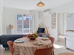 A dining area with a round table, chairs, a bowl of fruit, and a television in the background. - Les Cariatides 8 by Interhome (Ile d'Oléron)