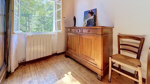 Ein Zimmer mit Holzboden, Heizkörper, einem fensterbelaubten Fenster, einem Holzschrank und einem Stuhl.