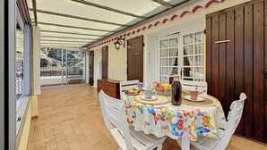 Ein überdachter Patio-Bereich mit Tisch, der für Tee gedeckt ist, umgeben von weißen Stühlen und Blick auf einen Garten durch eine Glastür.