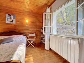 A cozy room with a wooden ceiling, a bed with a patterned bedspread, a director's chair, a bookshelf, and a window with a view of trees. - La Bergerie de Portes by Interhome (Portes)