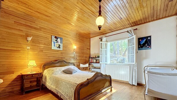 Ein Schlafzimmer mit Holzwandverkleidung, einem Bett, Nachttischlampen, einem Fenster mit Blick auf Bäume und einem weißen Heizkörper.