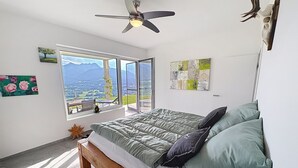 A bedroom with a bed, a ceiling fan, a large window with a mountain view, and artwork on the walls. - Karawanken Panorama by Interhome (Ludmannsdorf)