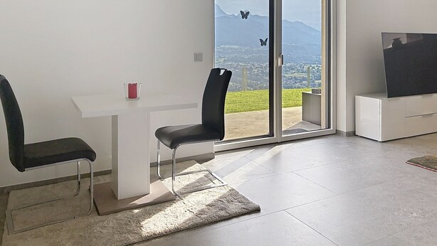 A modern living room with a sliding glass door leading to a balcony, a flat-screen TV, and a minimalist furniture arrangement. - Karawanken Panorama by Interhome (Ludmannsdorf)