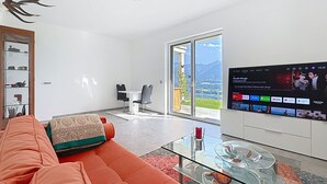 A modern living room with a glass coffee table, a sofa, and a flat-screen TV mounted on the wall. - Karawanken Panorama by Interhome (Ludmannsdorf)