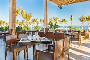 An outdoor dining area with tables set for two, surrounded by palm trees and overlooking the sea. - New Year 2BR 3BA on the beach in Exclusive Villa La Valencia, Los Cabos (Villa La Valencia, Los Cabos)