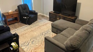 Un salon comprenant un canapé, un fauteuil inclinable, un téléviseur posé sur un meuble en bois, un tapis à motifs et une petite table sur laquelle se trouve une boîte de mouchoirs.