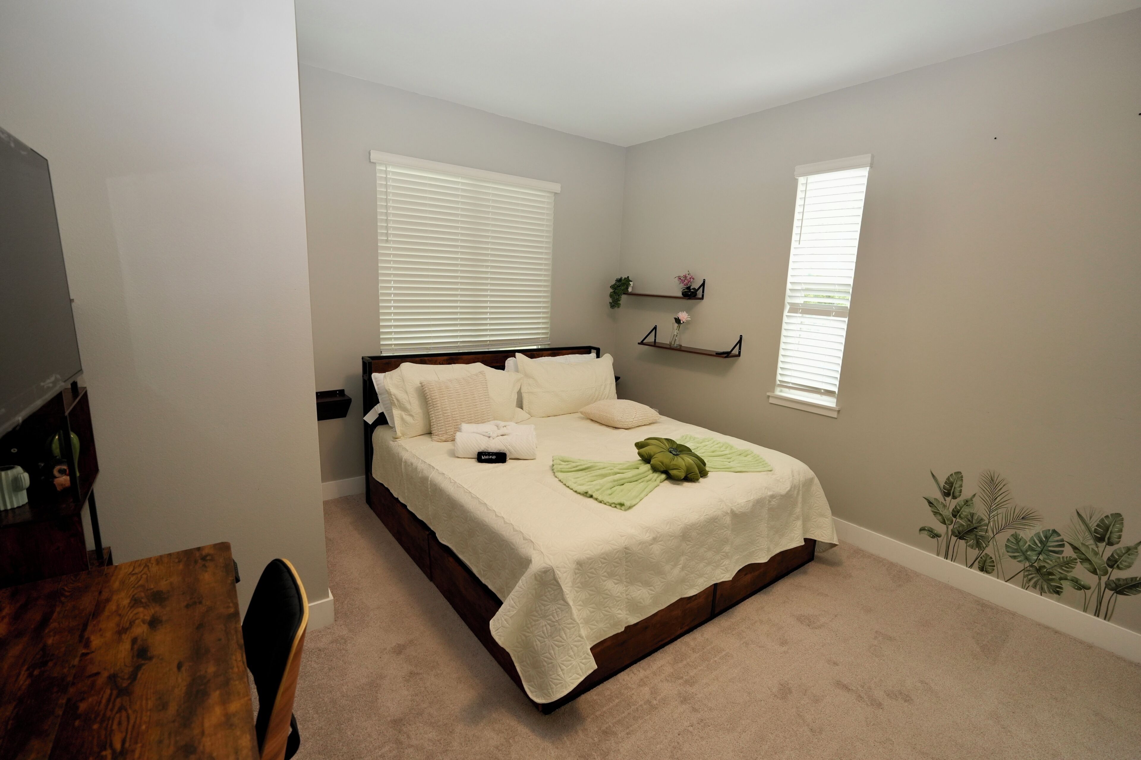 A bedroom with a bed, a wooden desk, and a chair. There are two windows with blinds, a shelf with decorative items, and a wall decal.