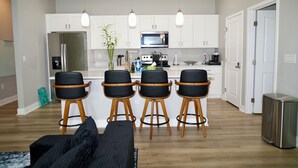 Una cucina moderna con mobili bianchi, un frigorifero in acciaio inox, un forno a microonde e una macchina per il caffè. Sono presenti quattro sgabelli da bar neri con gambe in legno e un divano nero con una coperta decorata.