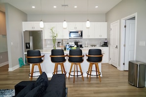 A modern kitchen with white cabinets, a stainless steel refrigerator, a microwave, and a coffee maker. There are four black bar stools with wooden legs and a black sofa with a patterned throw. - Orlando Escape • Near Disney • 5 Bedrooms (Orlando)