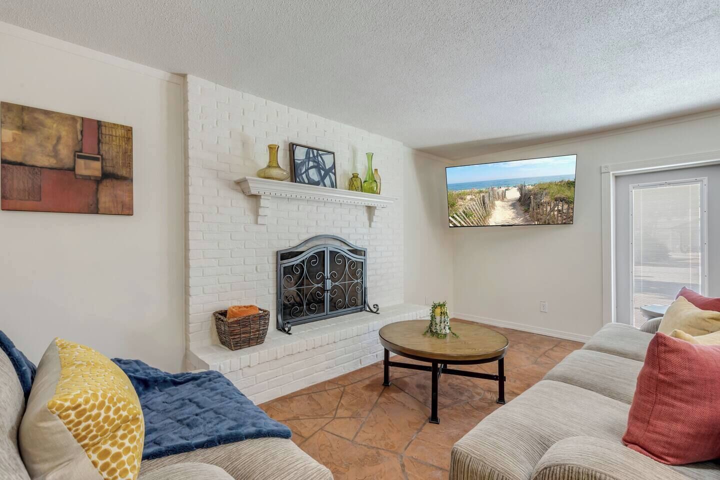 A living room with a fireplace, a sofa, a coffee table, and a view of the beach through a window.