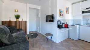 Un ambiente compatto con un divano, un piccolo tavolino rotondo, una TV, angolo cottura e una porta che conduce in un'altra stanza.