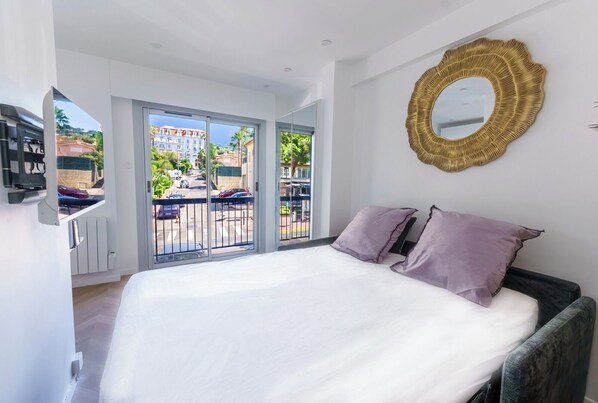 A bedroom with a large bed, a balcony with a view of buildings and greenery, and a decorative mirror on the wall. - Premium Studio • Antibes Street • Beach: 5 min (Cannes)