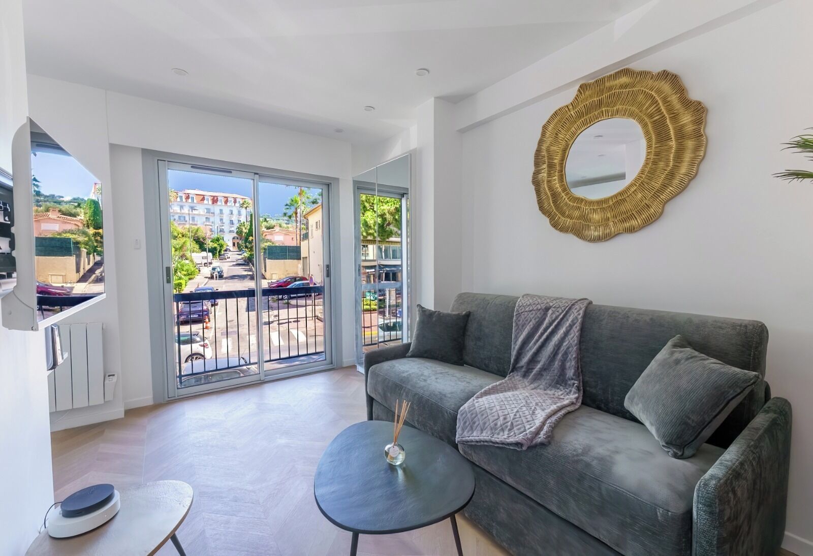 Een moderne woonkamer met een grijze bank, een klein rond tafeltje en een groot raam met uitzicht op een straat en gebouwen.