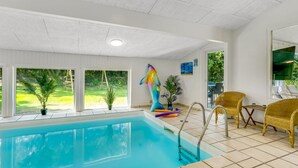 Uma piscina interior com janelas amplas, uma escultura colorida, cadeiras de vime e vista para o exterior com vegetação.