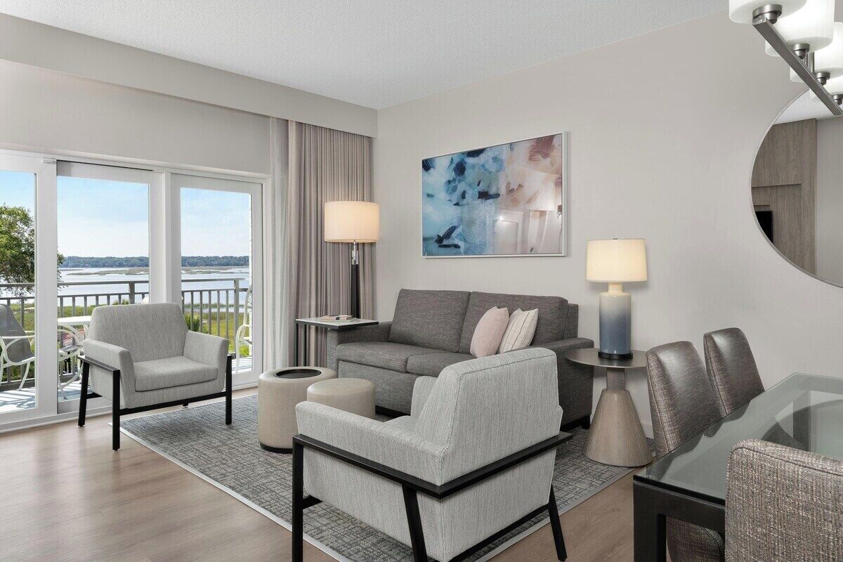 Une chambre d’hôtel moderne avec un balcon, un canapé gris, une table à manger en verre et un grand tableau abstrait.