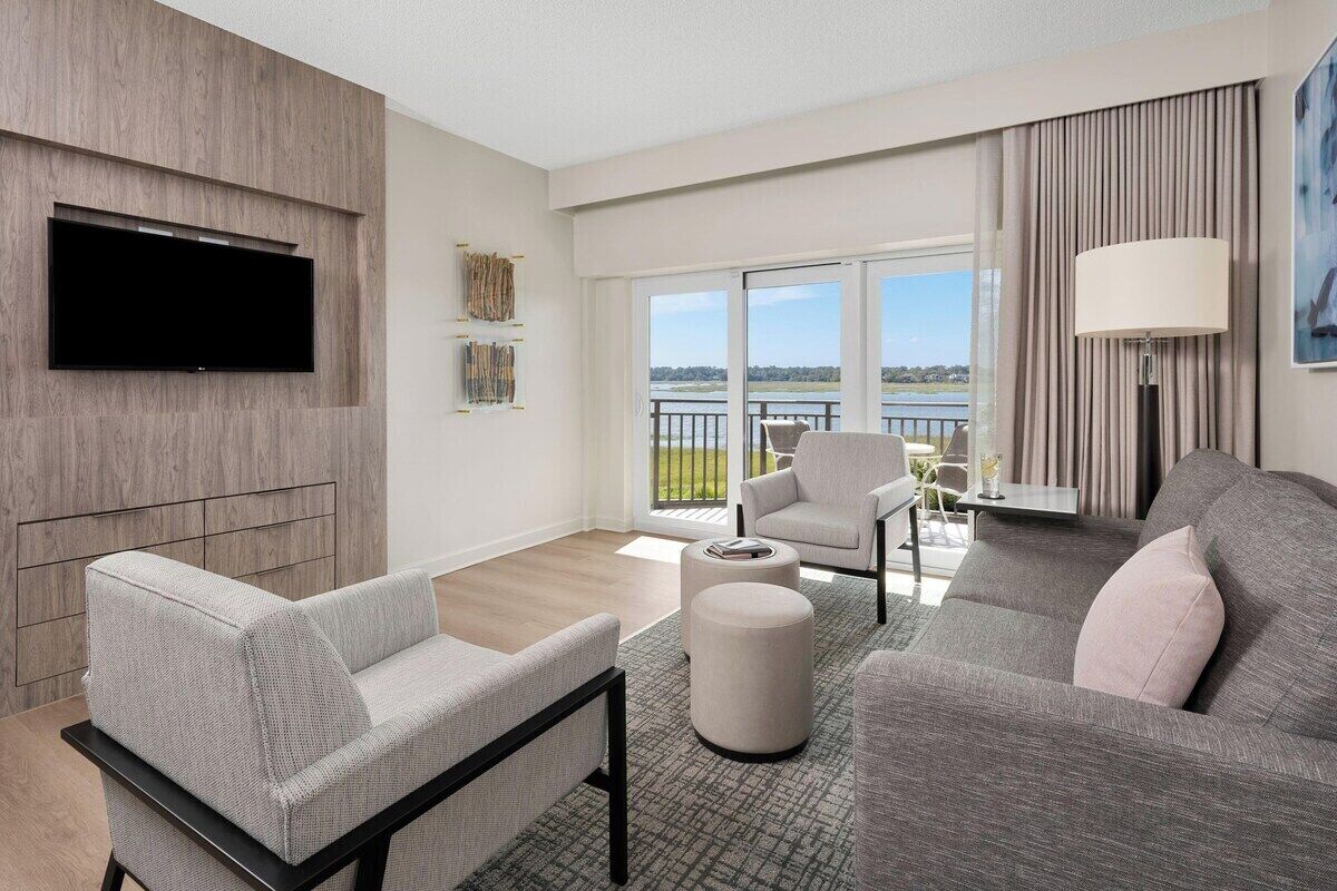 Ein modernes Wohnzimmer mit einem Flachbildfernseher, einem grauen Sofa, einem weißen Sessel und einem großen Fenster mit Blick ins Freie.