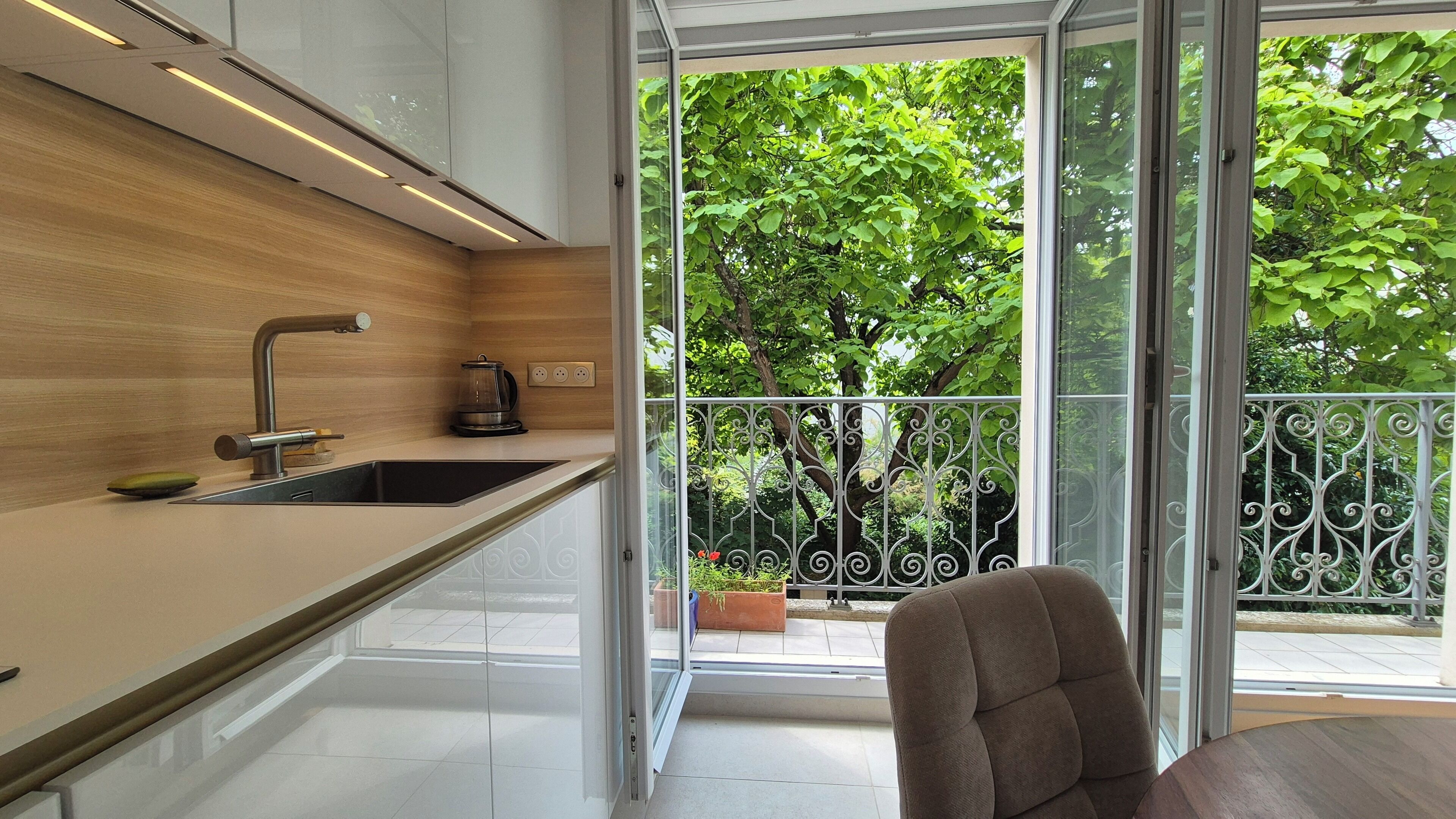 Une cuisine moderne dotée d’un évier encastré, d’une machine à café et offrant une vue sur un balcon avec une rambarde en métal et de la verdure.
