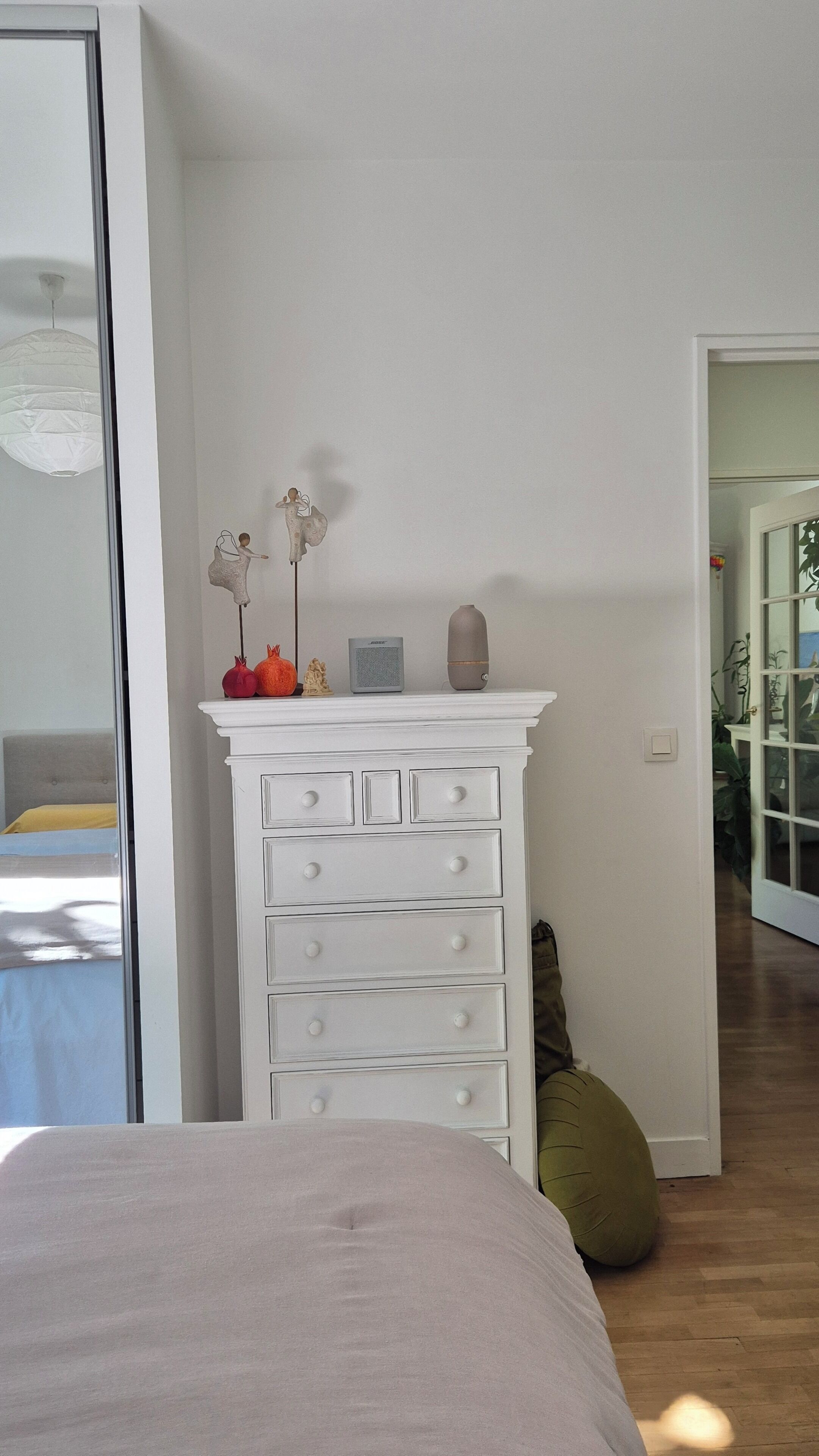 A bedroom with a bed, a dresser with drawers, a potted plant, and a door leading to another room.