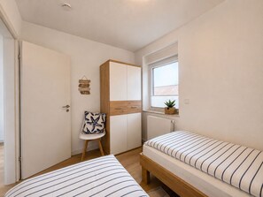 Ein Schlafzimmer mit zwei Betten, einem Kleiderschrank, einem Sessel, einem Fenster mit Pflanze und einem Schild an der Wand.
