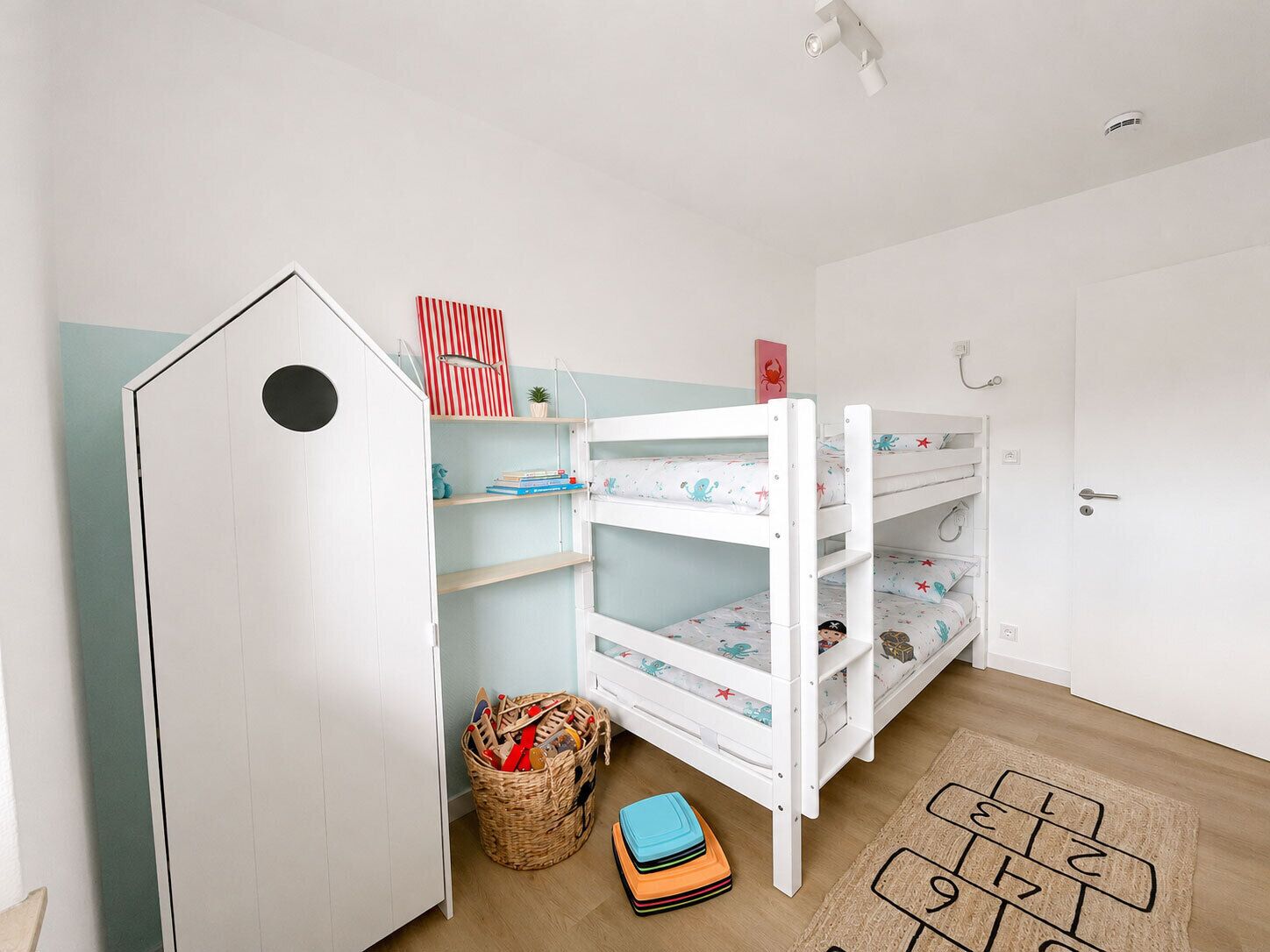 En børne-køjeseng med hvid ramme og mønstret sengetøj, en lille hylde med bøger og legetøj, en kurv med trælegetøj og en hopscotch- måtte på gulvet.