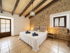 Ein Schlafzimmer mit Steinwand, einem Bett mit weißer Bettwäsche, einer Holzbalkendecke und einem Fenster mit Vorhängen.
