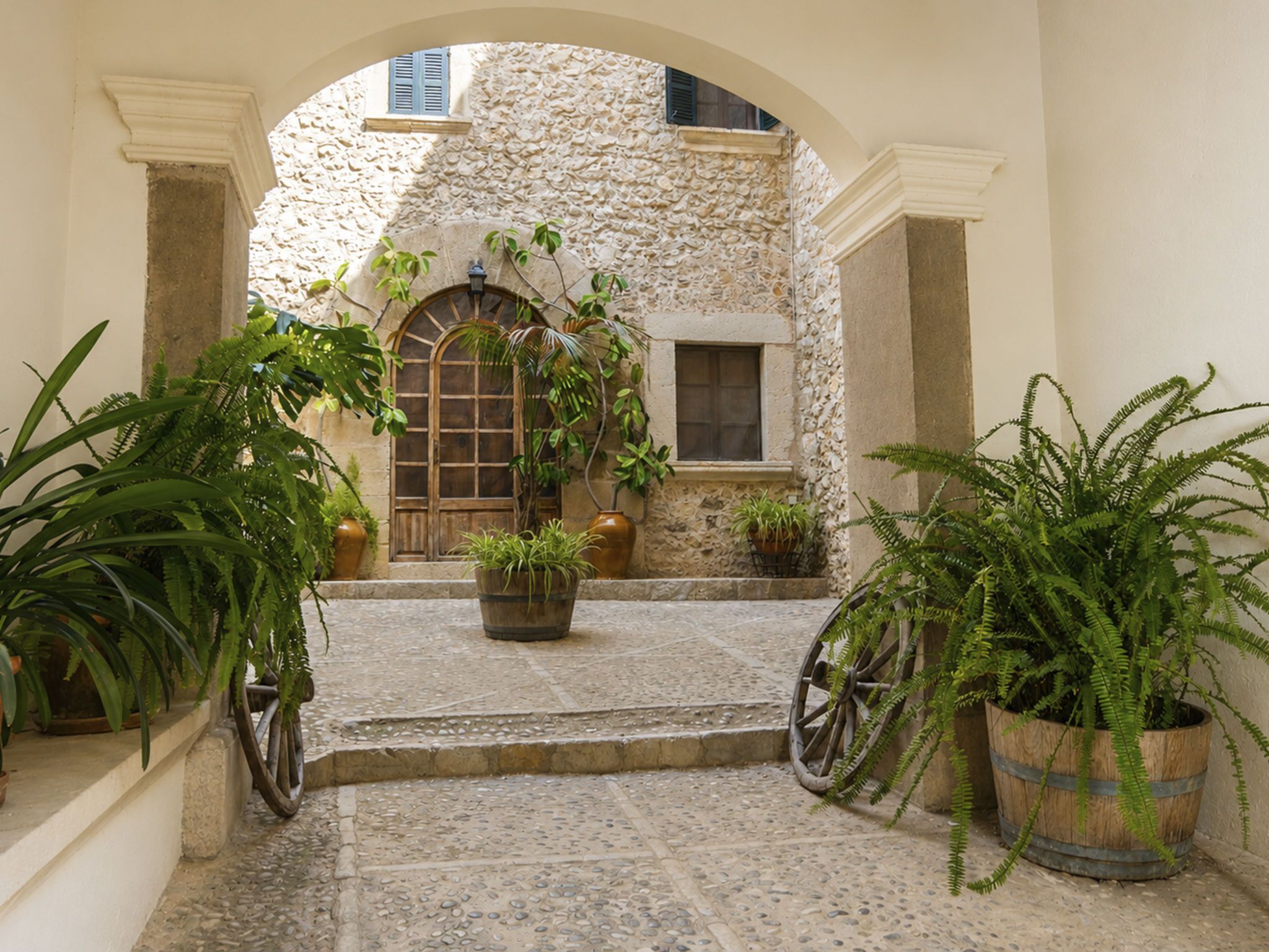 Une entrée en arc donnant sur un bâtiment à la façade en pierre, agrémentée de plantes en pot et d’un vieux chariot en bois.