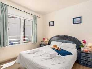 Ein ordentlich bezogenes Bett mit weißen Bettwäsche, einer blauen Decke und einem Hut daneben. Es gibt zwei Nachttische mit Lampen und Dekoration.