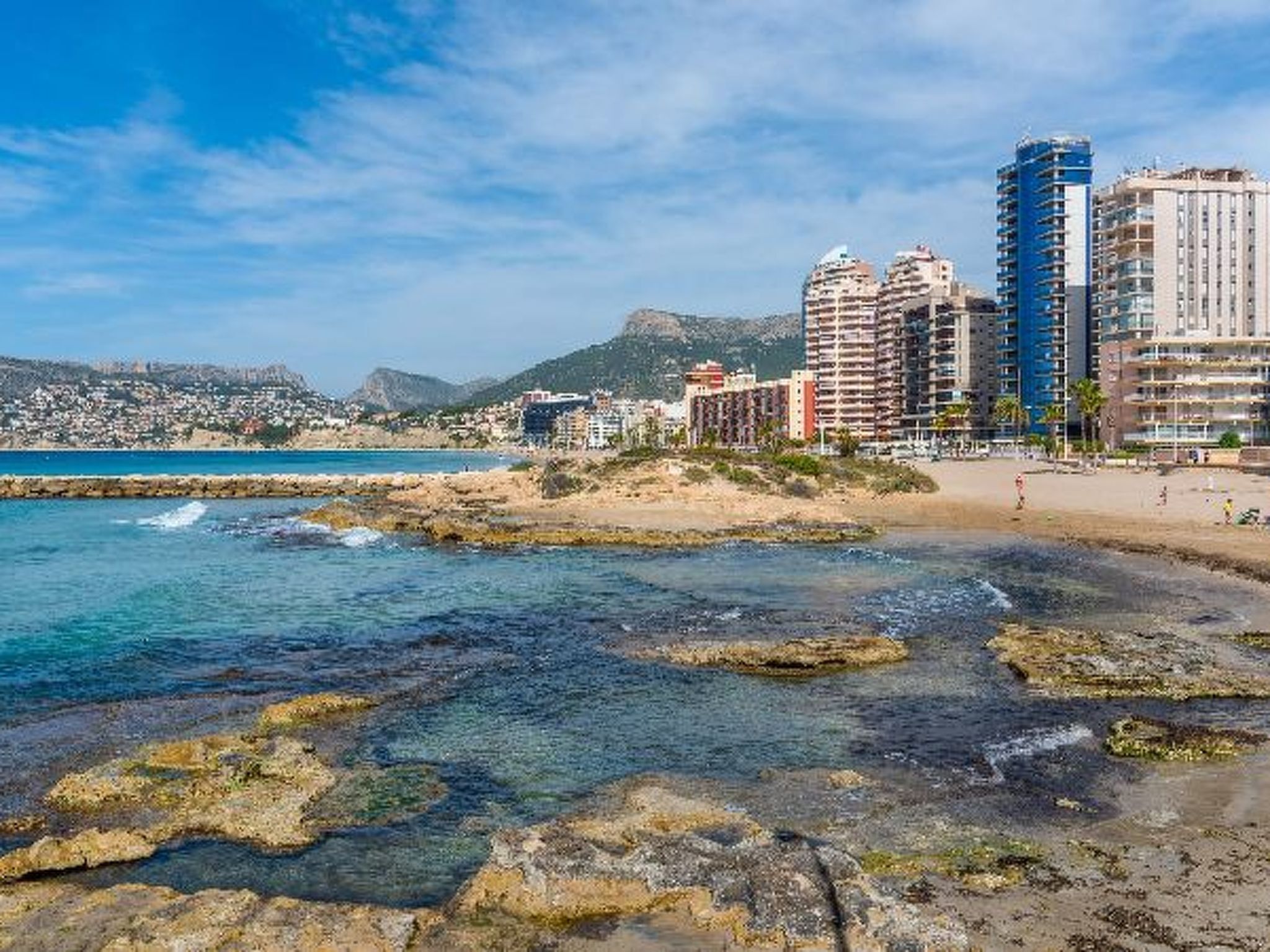 A coastal cityscape with a beach, rocky shoreline, and high-rise buildings.