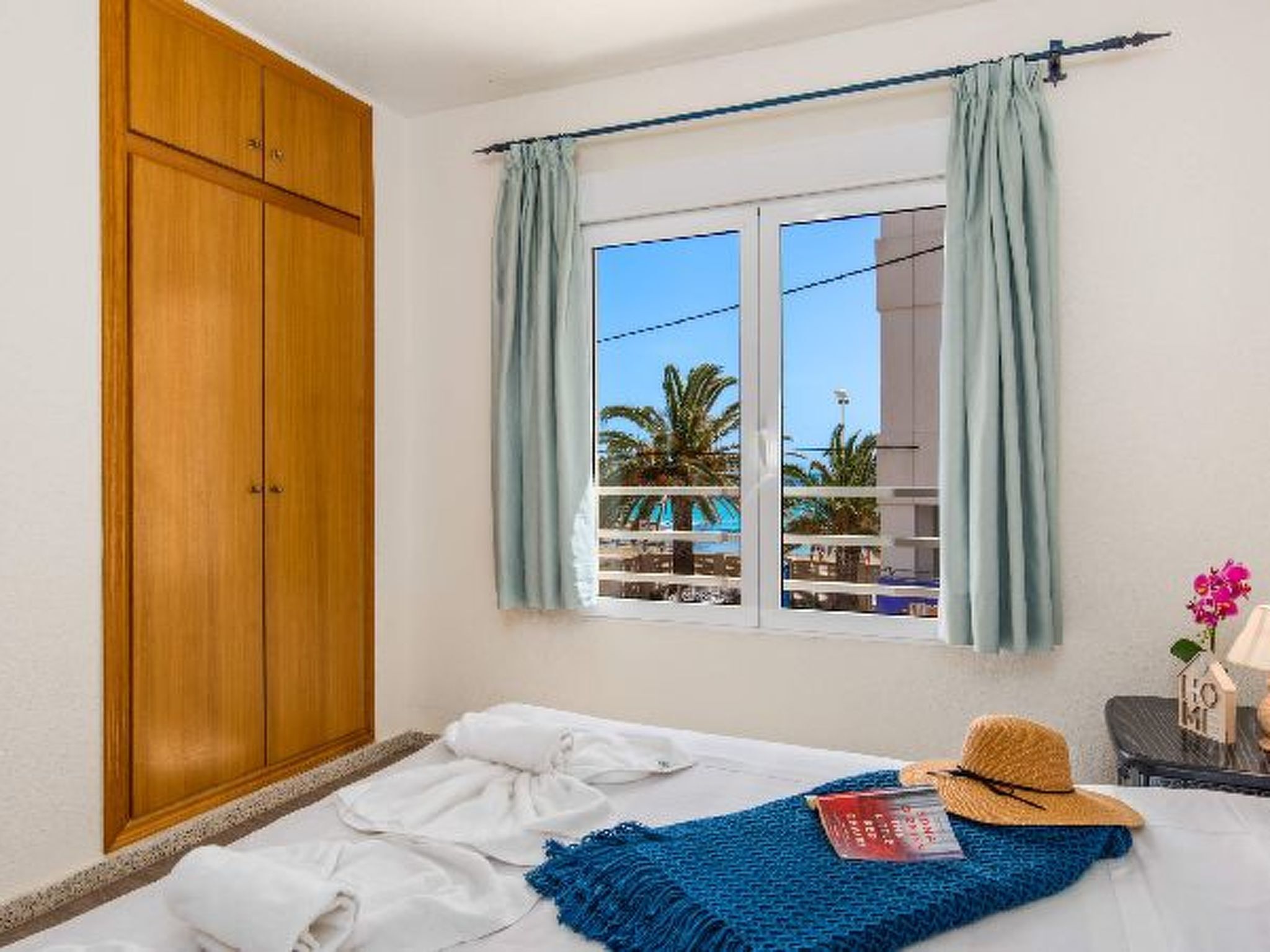 Un dormitorio con una cama, un armario de madera, una ventana con cortinas, vista a palmeras y una pequeña mesa con un sombrero y un libro.