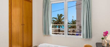 Un dormitorio con una cama, un armario de madera, una ventana con cortinas, vista a palmeras y una pequeña mesa con un sombrero y un libro.