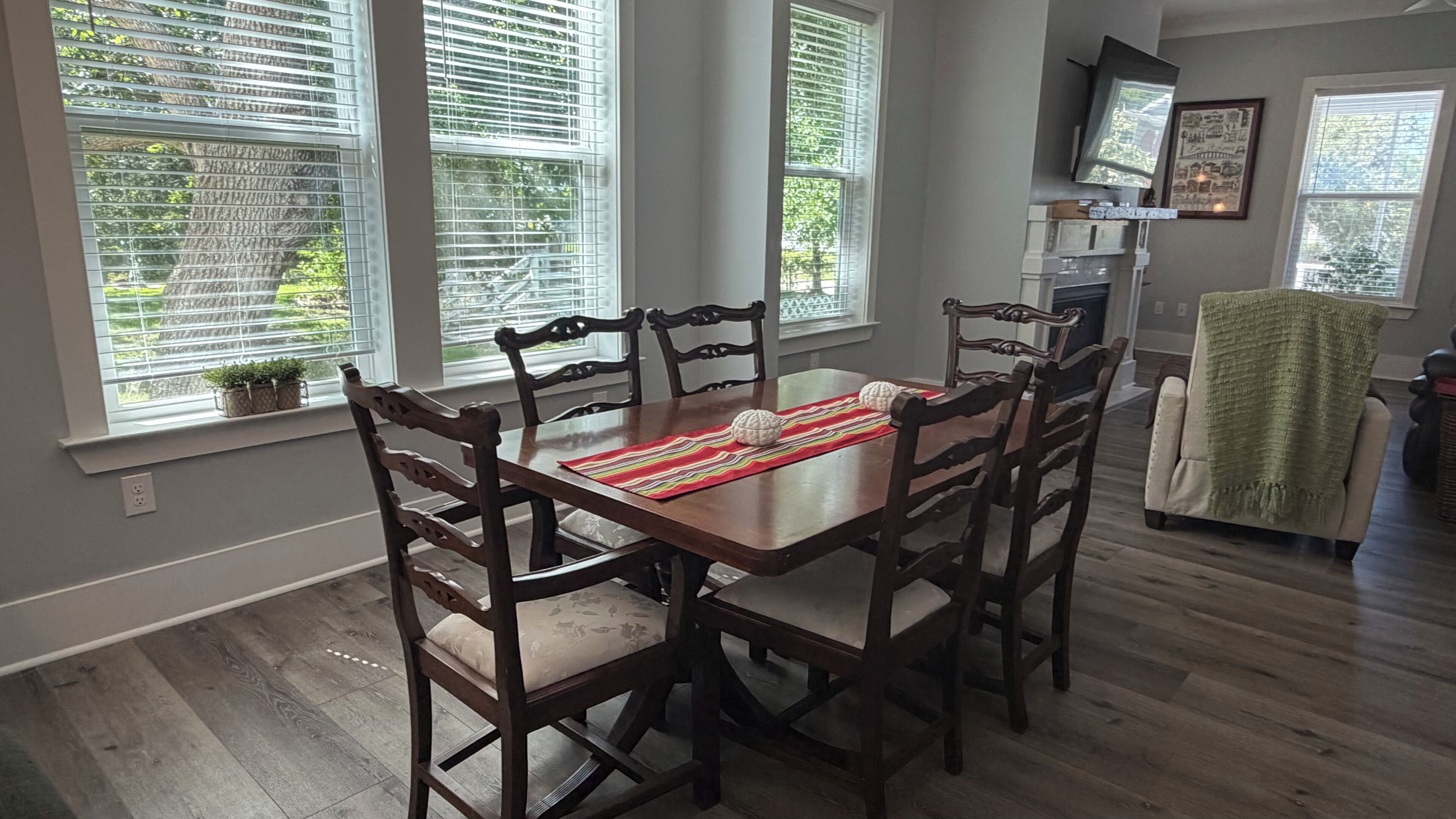 A dining area with a wooden table and chairs, large windows with blinds, and a television mounted on the wall.