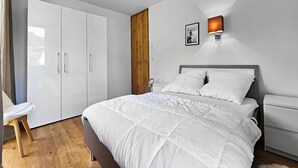 Un dormitori amb un llit, un armari, una tauleta de nit, una làmpada penjada a la paret i una porta de fusta.