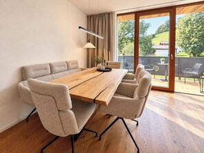 A modern dining area with a wooden table, beige upholstered chairs, and a view of a green landscape through large sliding glass doors. - Das Weidach by Interhome (Brixen im Thale)