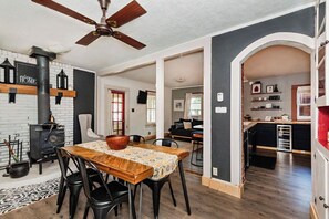 A dining area with a wooden table, chairs, a ceiling fan, a brick stove, and a kitchen visible through an archway.