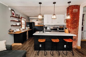 Una cuina amb una illa central, tamborets de bar, llums penjants i una paret decorativa de maó.