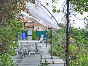 Eine sonnige Terrasse mit Tisch und Stühlen, umgeben von viel Grün und einem Haus im Hintergrund.