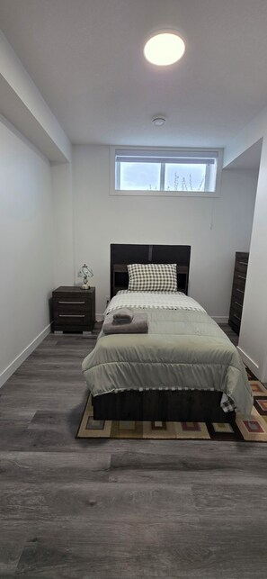 Ein Schlafzimmer mit Bett, Nachttisch, Kommode, Fenster und Deckenleuchte.