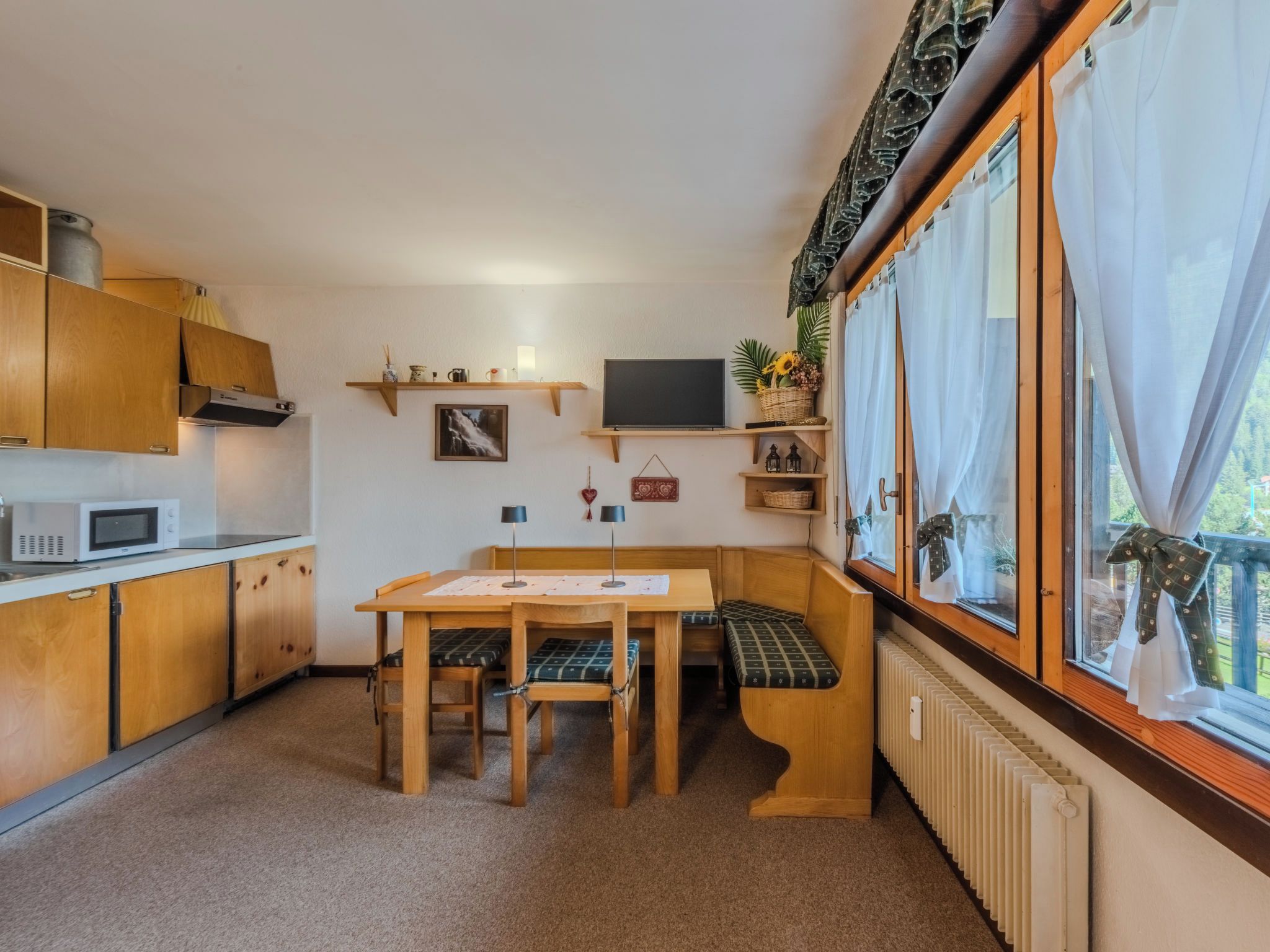 Een compacte keuken met houten kastjes, een magnetron, een eettafel met bankjes en een raam met gordijnen.