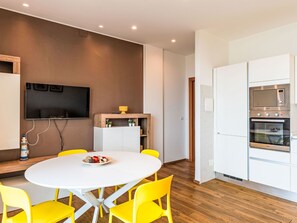Eine moderne Küche mit integrierten Geräten, eine Essecke mit einem runden Tisch und gelben Stühlen sowie ein flachbildfernseher an der Wand.