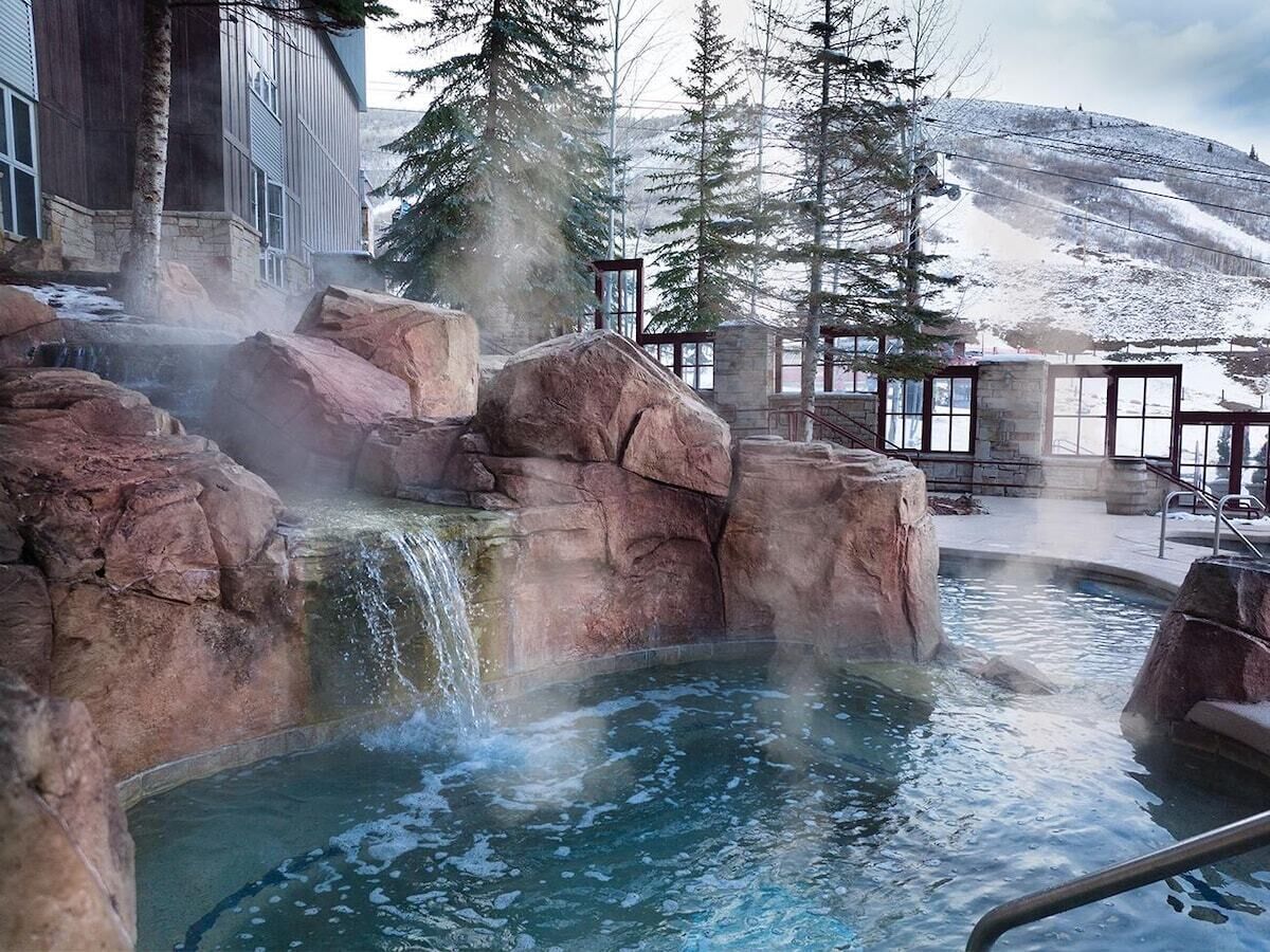 Une zone de sources chaudes avec une cascade, entourée de grands rochers et d’arbres, avec un bâtiment visible au loin.