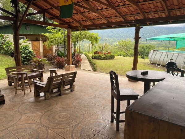 An outdoor patio with wooden furniture, a covered area with a green roof, and a view of a mountainous landscape.