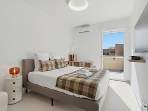 Ein modernes Schlafzimmer mit einem Bett, Nachttischen, einem Fernseher und Blick auf einen Balkon.