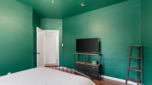 Un dormitorio con una cama, un televisor sobre un mueble, una puerta y una estantería con una cesta.