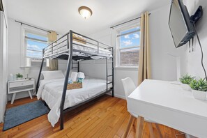 Een slaapkamer met een stapelbed, bureau en stoel, een aan de muur bevestigde televisie en een raam met gordijnen.